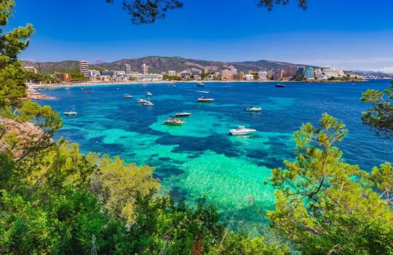 Playa con aguas turquesas y barcos, edificios al fondo y árboles en primer plano.