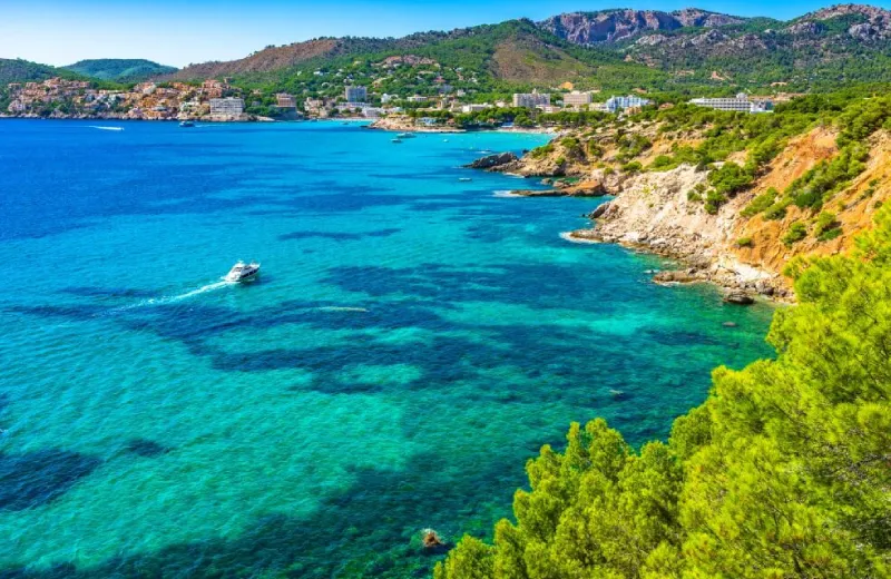 Costa rocosa con agua turquesa, vegetación y edificaciones al fondo bajo un cielo despejado.