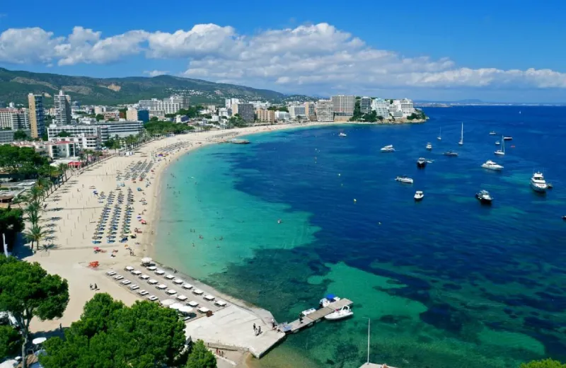 Vista aérea de una playa con sombrillas, edificios al fondo y barcos en el mar azul.