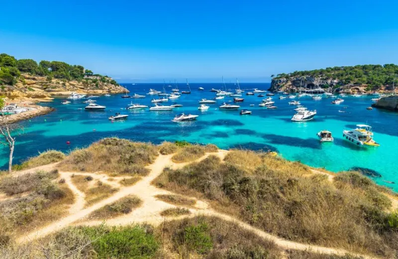 Bahía con agua turquesa, barcos anclados y orilla cubierta de vegetación. Cielo despejado.