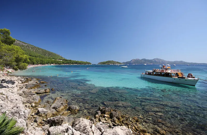 Barco en agua turquesa cerca de la costa rocosa, con colinas y cielo despejado al fondo.