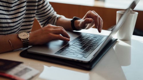 Persona tecleando en una laptop sobre una mesa con taza.