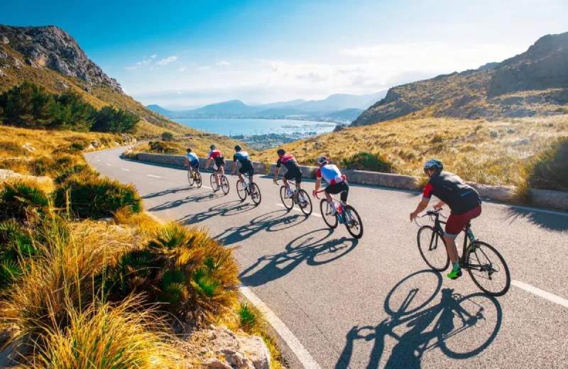 Ciclismo en grupo por carretera montañosa con vistas al mar y cielo despejado.