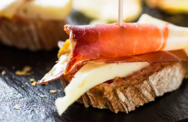 Tosta de pan con queso y jamón serrano enrollado, presentada sobre una superficie oscura.