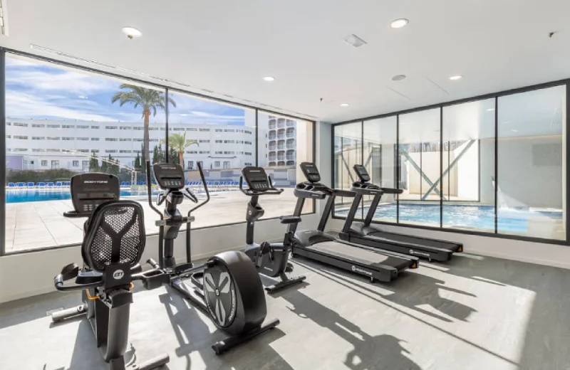 Gimnasio con bicicletas, caminadoras y vista a la piscina exterior a través de un ventanal.
