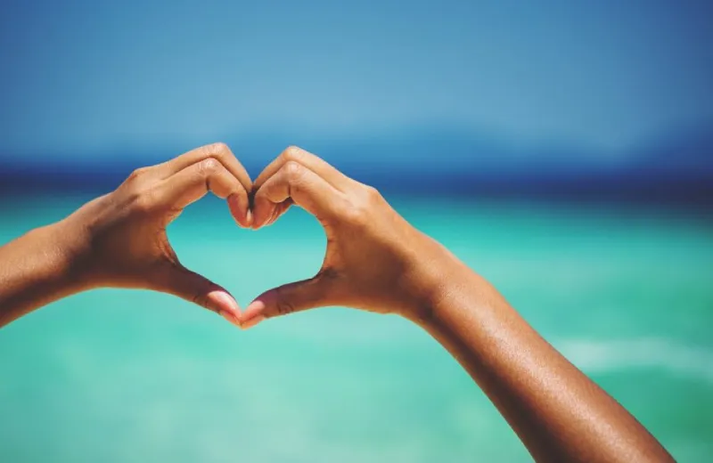 Manos formando un corazón frente a un fondo de mar azul claro.