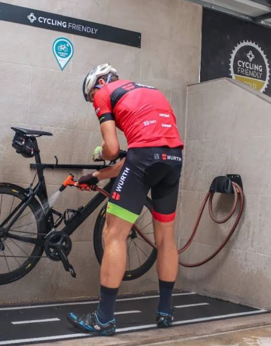 Ciclista limpiando su bicicleta en una estación "Cycling Friendly" con manguera y equipamiento.