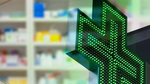 Cruz de farmacia verde iluminada, con estantes de medicamentos desenfocados al fondo.