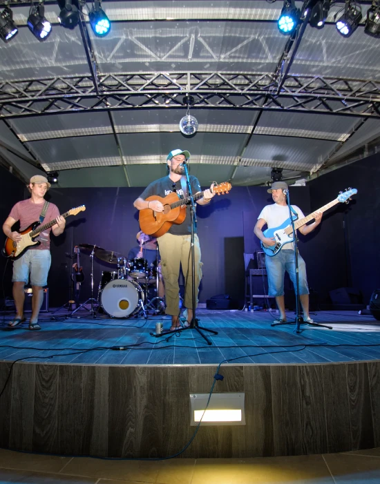 Banda de cuatro músicos tocando en un escenario con luces azules y techo cubierto.