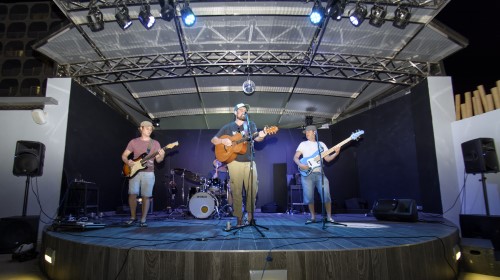 Banda tocando en un escenario al aire libre con luces, guitarras y batería.