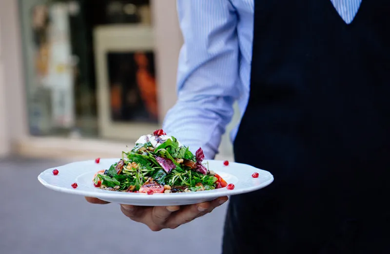 Persona con delantal sostiene un plato de ensalada con granada y hojas verdes.
