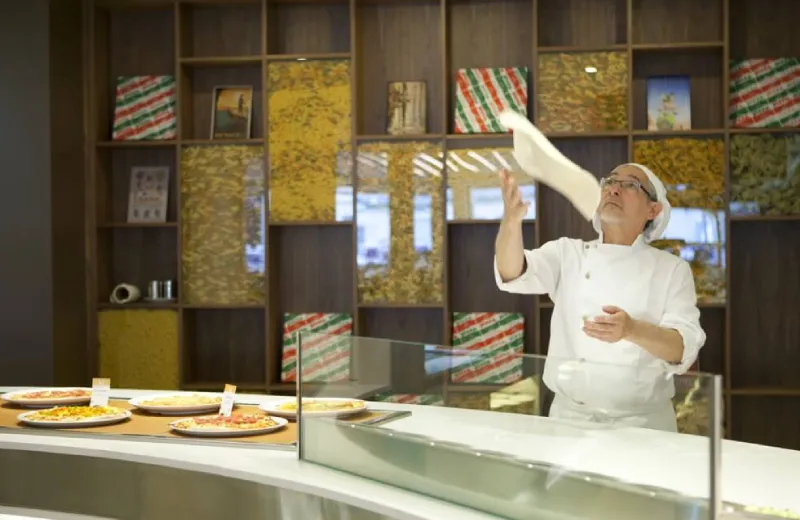 Persona en cocina lanzando masa de pizza al aire, con varias pizzas listas sobre el mostrador.
