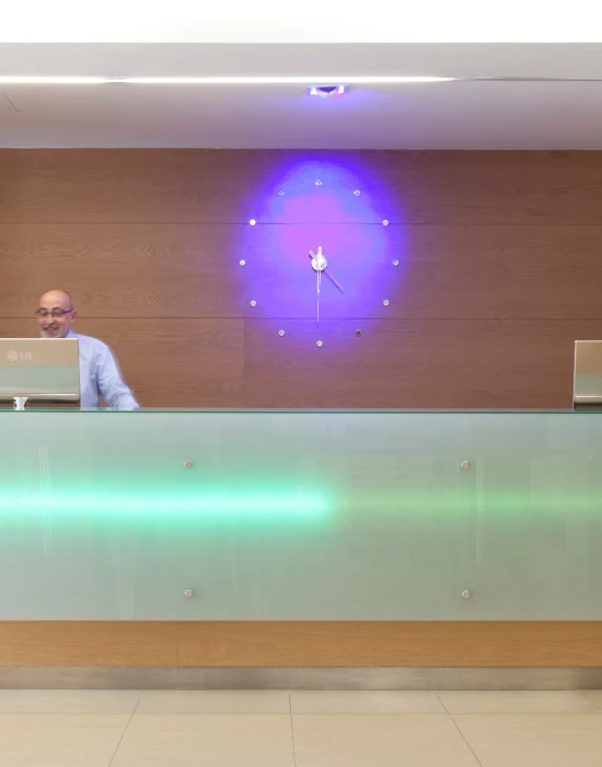 Recepción moderna con mostrador de cristal y reloj iluminado en la pared. Persona de pie detrás.