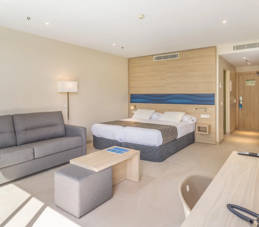 Habitación de hotel moderna con cama doble, sofá gris, mesa pequeña y decoración minimalista.