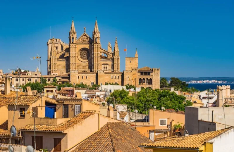 Vista de la Catedral de Mallorca sobre techos de la ciudad, con cielo azul y mar de fondo.