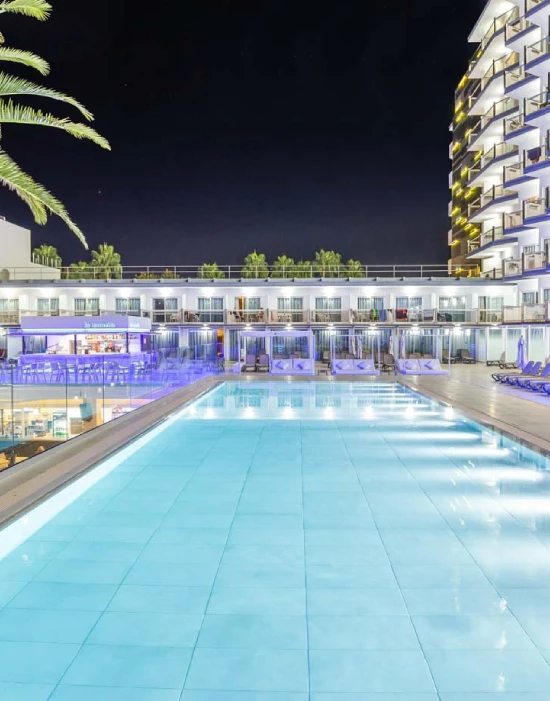 Piscina iluminada de noche en un hotel, rodeada de edificios y palmeras a la derecha.