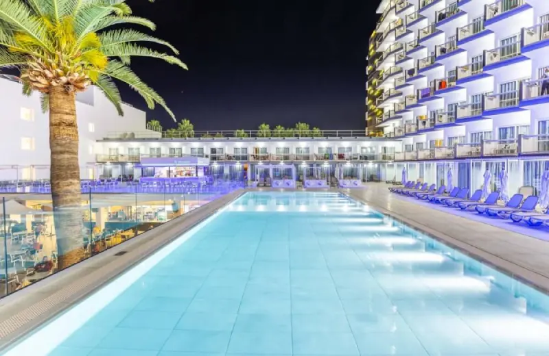 Hotel con piscina iluminada de noche, rodeado de palmeras y tumbonas.