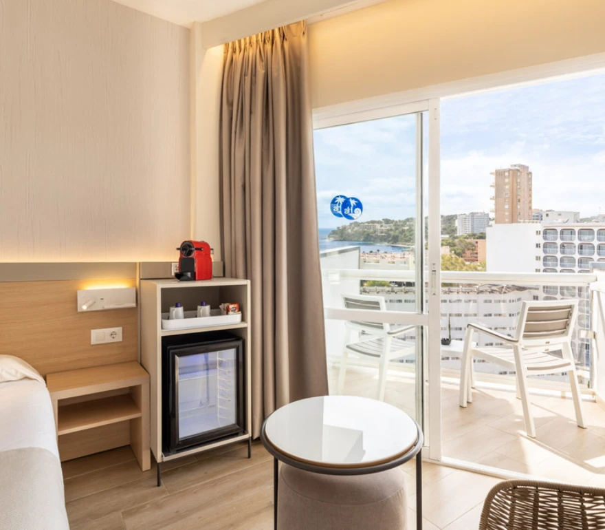 Habitación de hotel con balcón, cafetera roja y minibar, vista al mar y edificios.