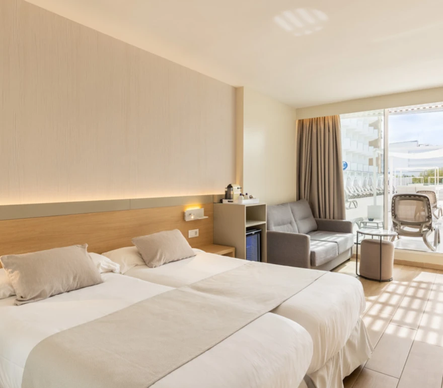 Habitación de hotel moderna con cama doble, sofá, y ventanal con salida a terraza con sillas.