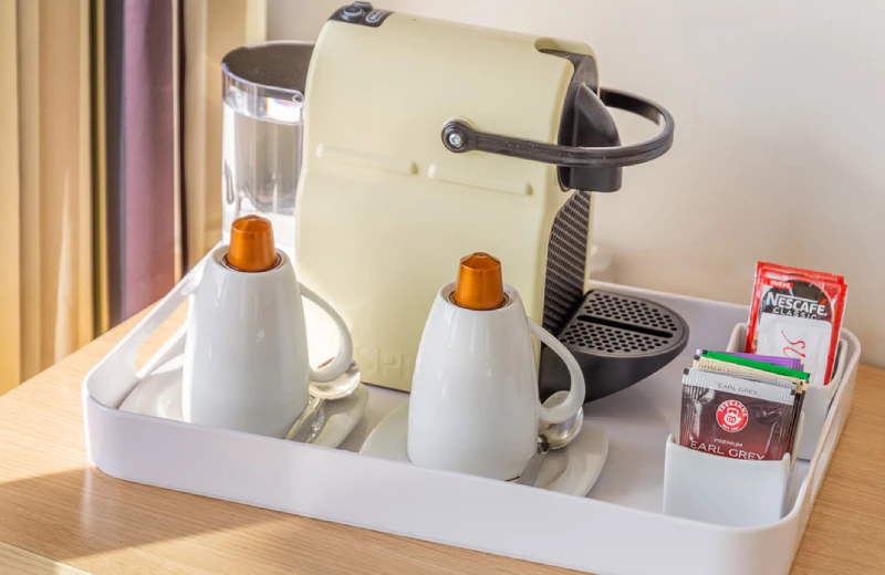 Máquina de café con cápsulas, tazas y sobres de té en una bandeja sobre una mesa de madera.