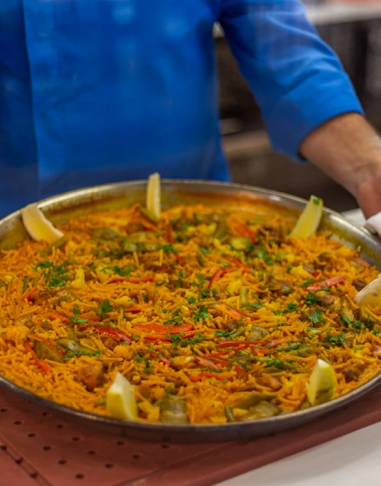 Persona sosteniendo una paellera con paella valenciana decorada con rodajas de limón.