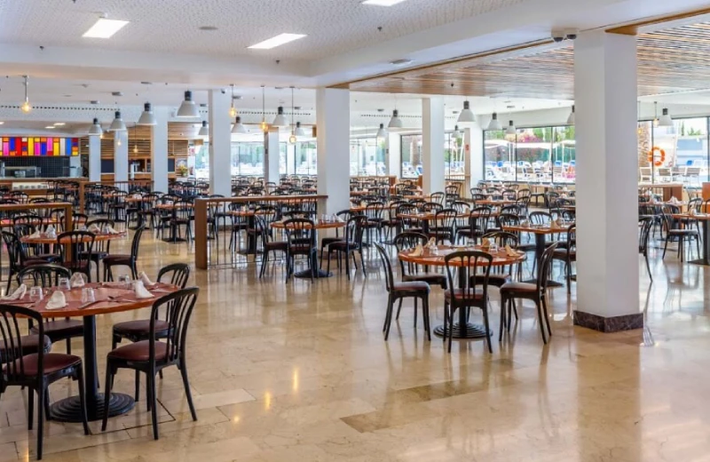 Un amplio comedor vacío con mesas redondas y sillas, iluminado por luz natural y lámparas colgantes.