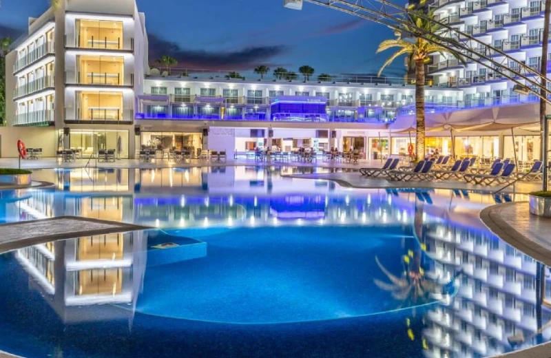 Hotel iluminado al anochecer con piscina reflejando las luces, palmeras y tumbonas.