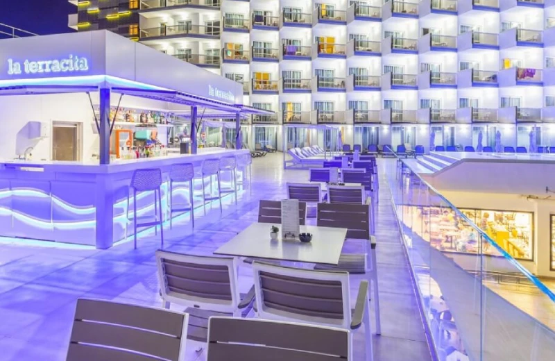 Bar al aire libre iluminado en terraza de hotel con mesas y sillas, frente a habitaciones iluminadas.