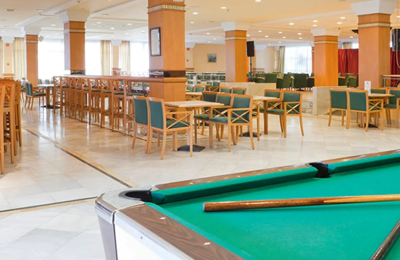 Mesa de billar en un amplio salón con mesas y sillas de madera y cojines verdes.