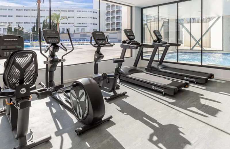 Gimnasio moderno con bicicletas estáticas y cintas de correr junto a una piscina iluminada.
