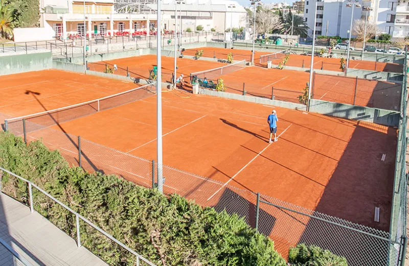 Vista de canchas de tenis de arcilla con jugadores practicando, rodeadas de edificios y jardines.
