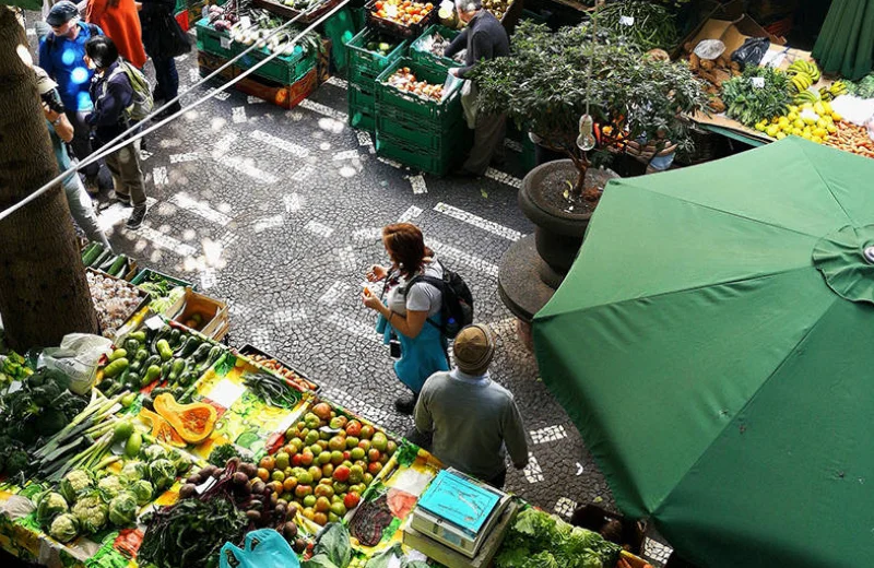 Vista aérea de un mercado al aire libre con frutas y verduras, gente caminando y una sombrilla verde.