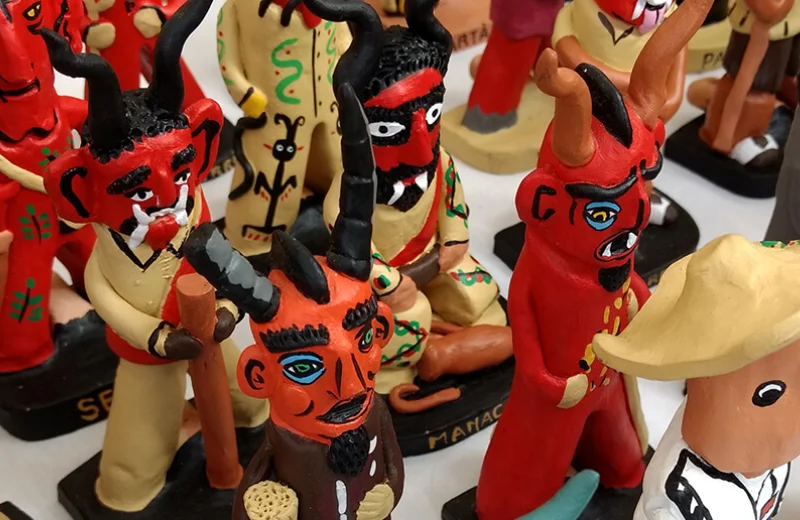 Figuras de diablos de cerámica pintadas de rojo con vestimenta colorida y cuernos negros.
