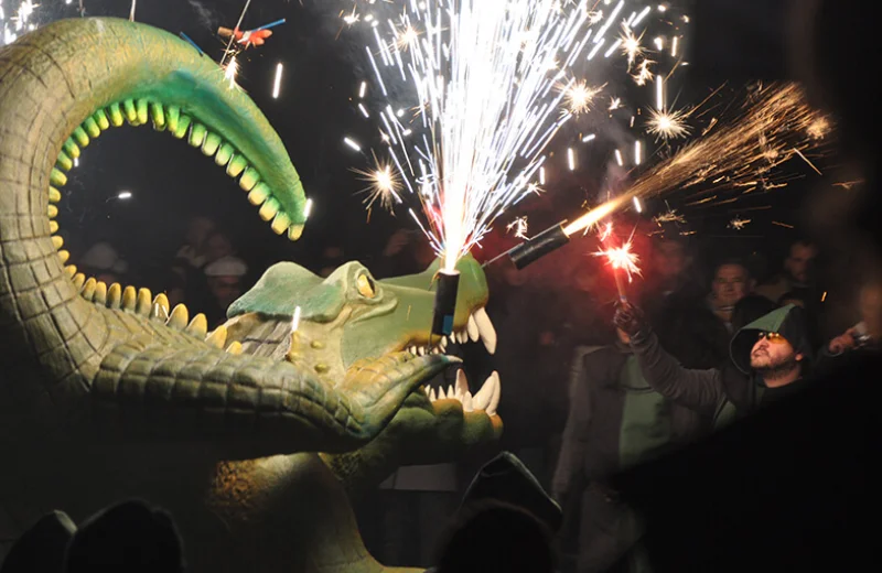 Escultura de cocodrilo con fuegos artificiales en su boca, rodeada de personas en la noche.
