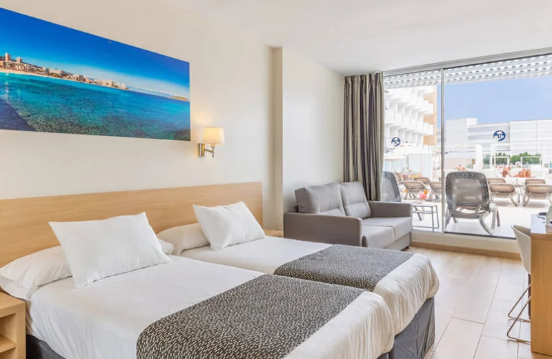 Habitación de hotel luminosa con dos camas, sofá, cuadro de playa y gran ventana con vistas al exterior.