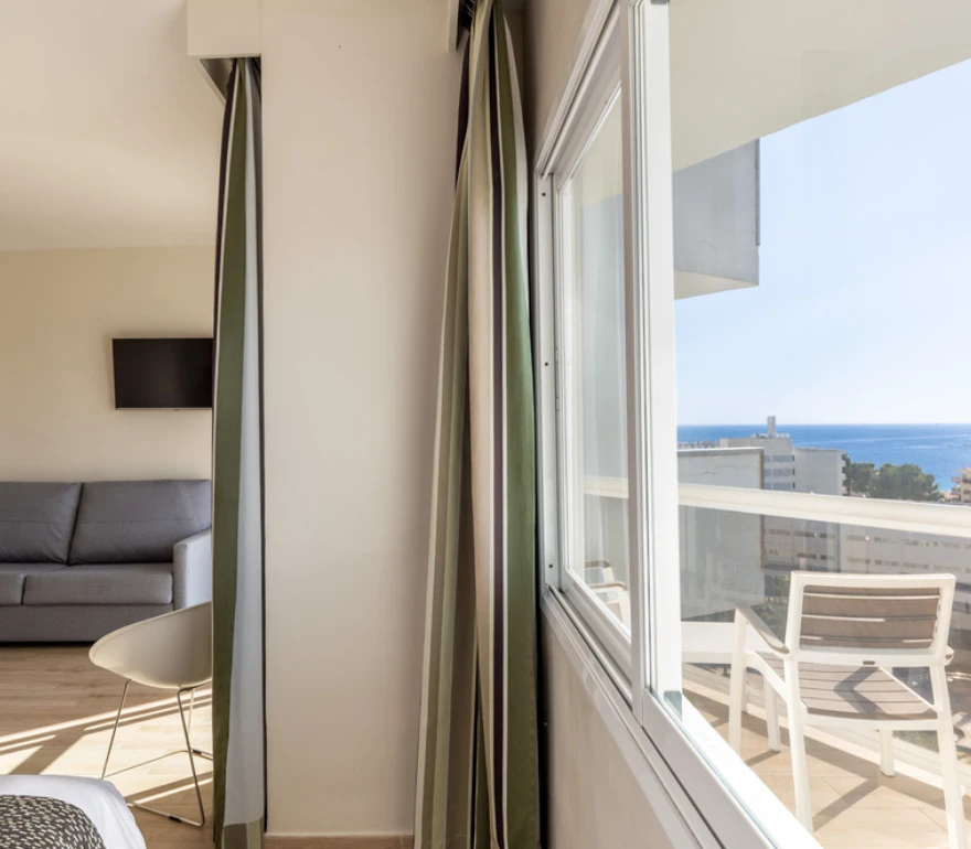 Sala luminosa con sofá, cortinas verdes y vista al mar desde un gran ventanal.