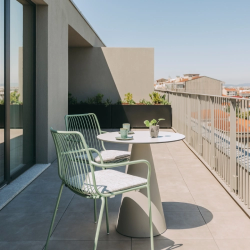 Two green chairs and a table on a balcony with a city view.