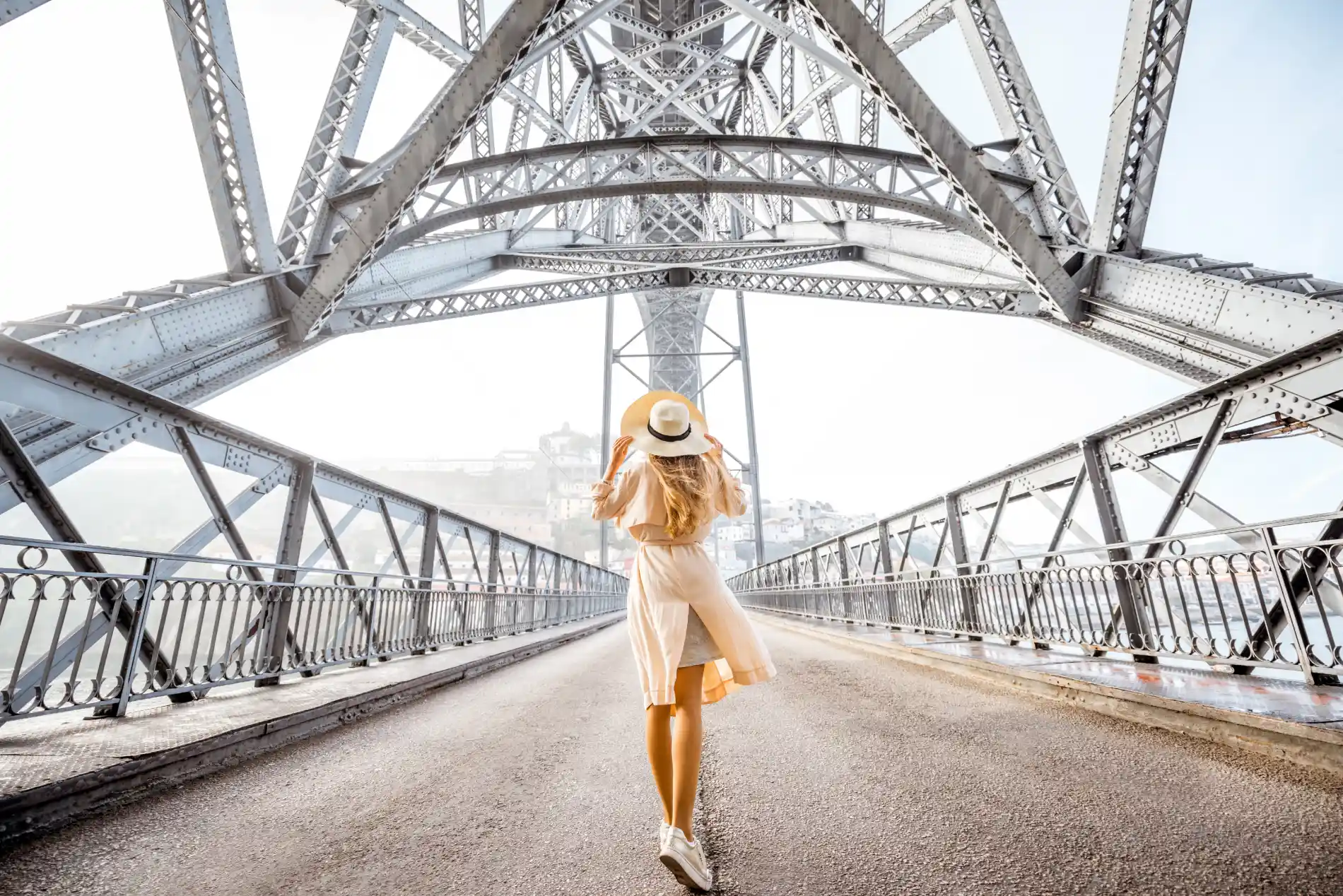 A woman in a dress and hat walks on a metal bridge holding onto her hat.