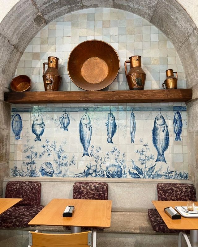 Ceramic wall with blue fish tiles, copper pots on top, wooden tables and chairs in foreground.