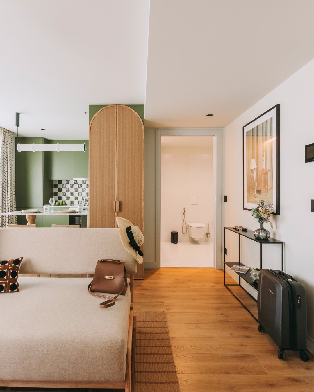 Modern apartment interior with sofa, kitchen, and hallway to a bathroom. Suitcase by the door.