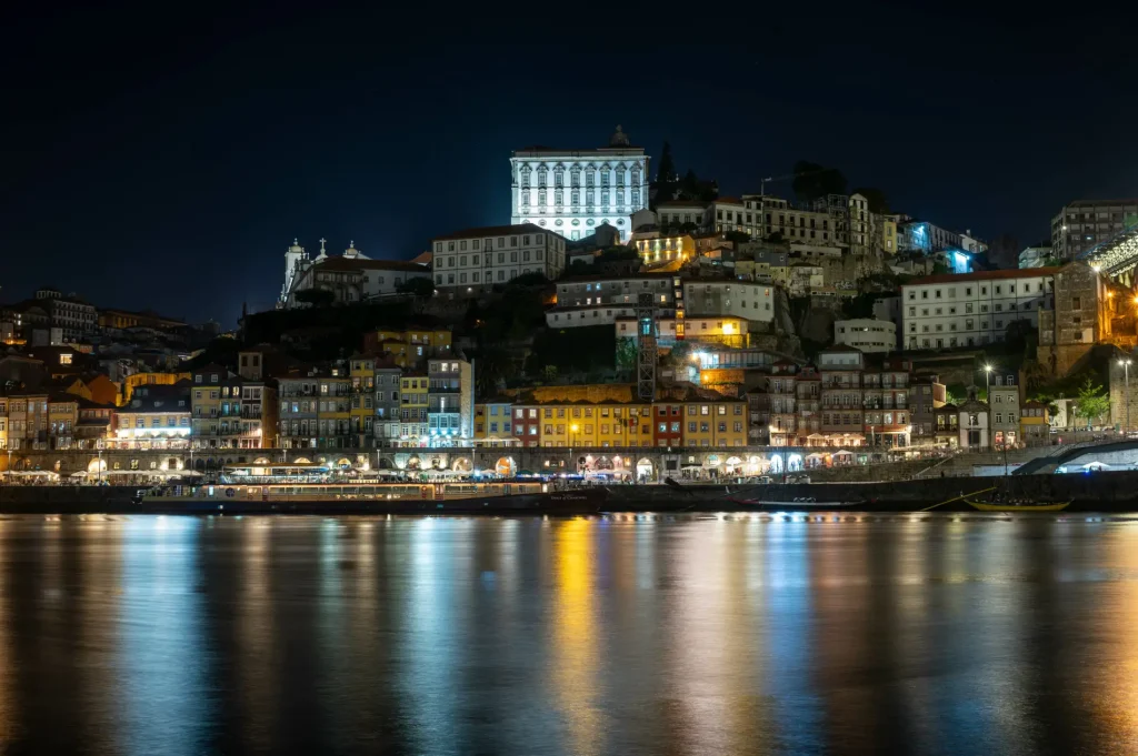 Cityscape at night with lit buildings reflecting on a calm river.