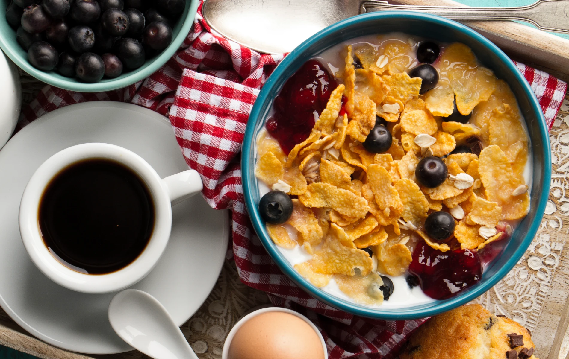 Breakfast tray with cereal and berries, coffee, a boiled egg, and a muffin on a checkered cloth.