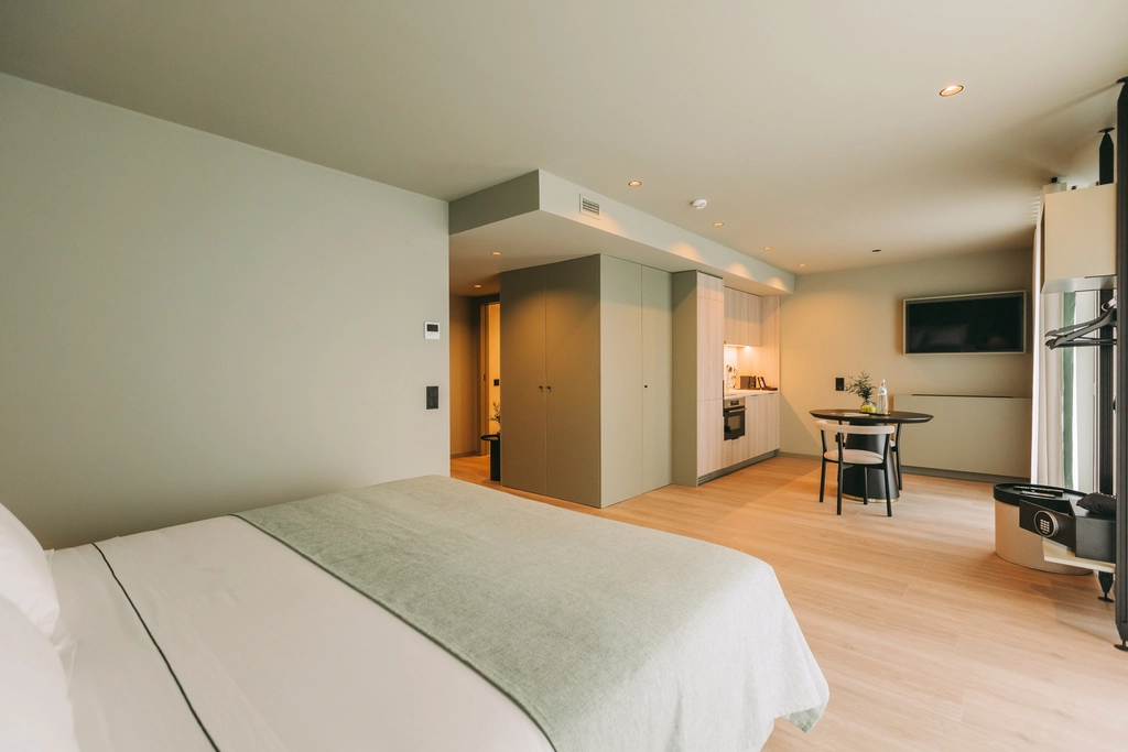 Modern hotel room with a bed, kitchenette, table, chairs, and TV. Light, minimalistic decor.