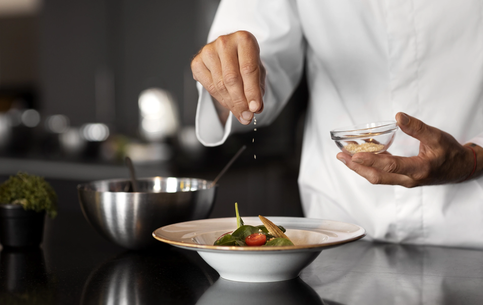 Chef sprinkling seasoning over a dish in a kitchen setting.