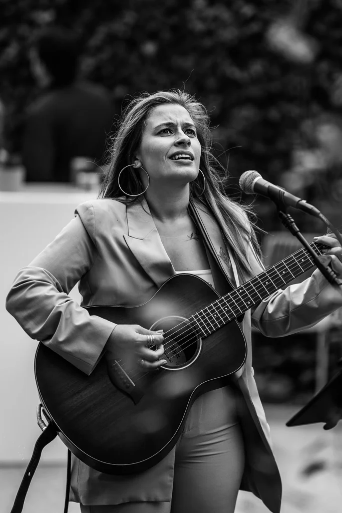 Woman singing and playing guitar on stage, wearing a blazer, with a microphone nearby.