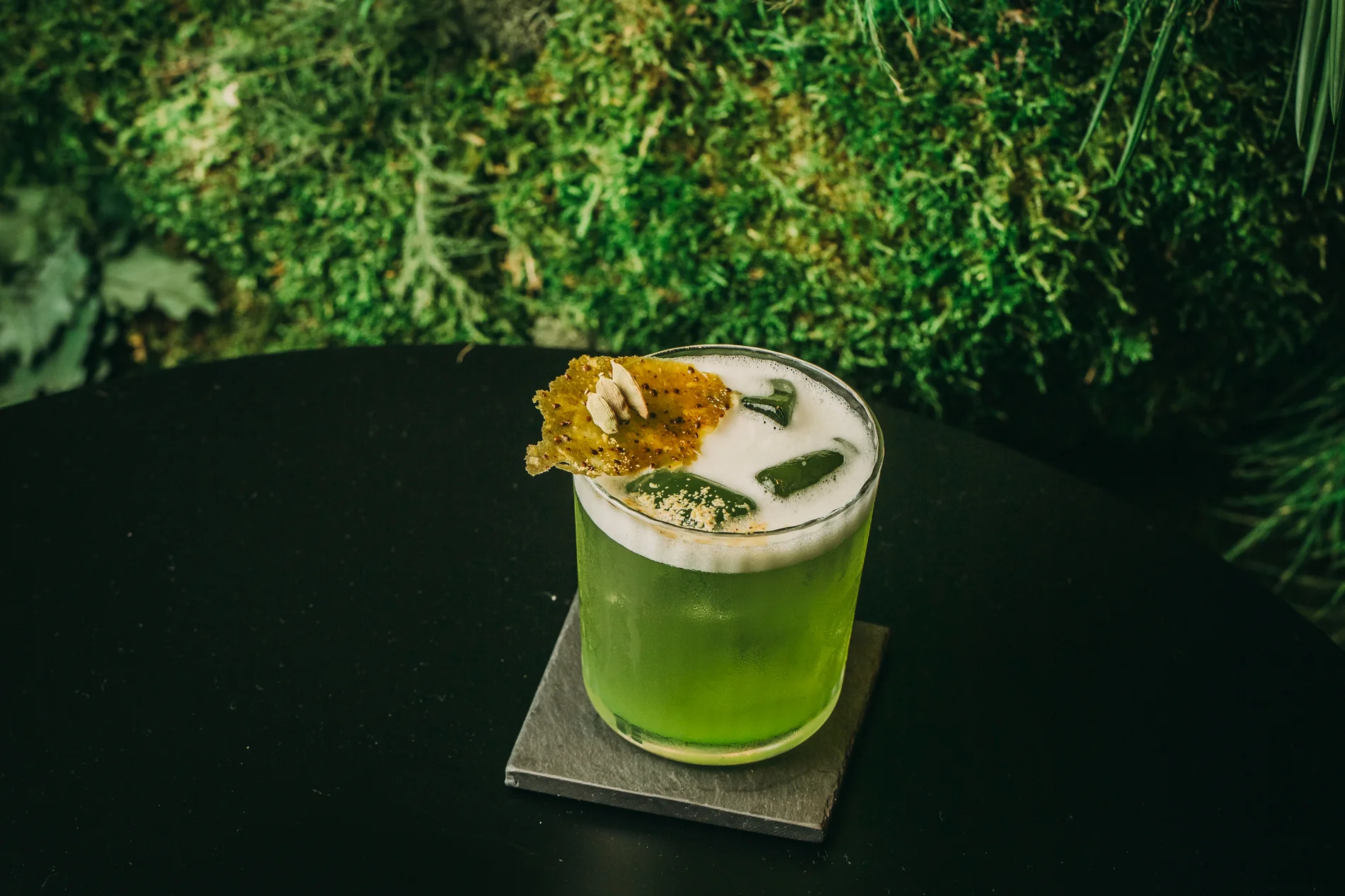 Green cocktail with frothy top on a coaster, garnished with a crispy chip and seeds.
