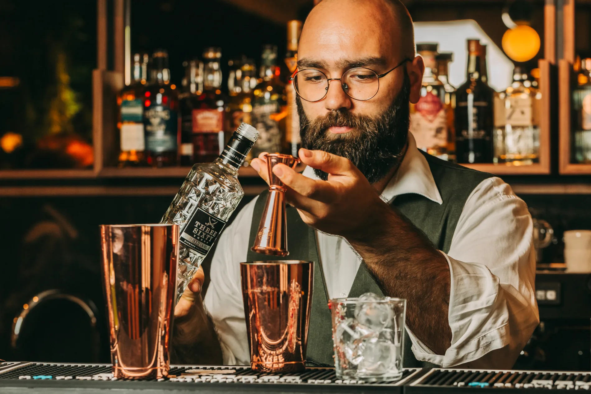 Bartender pouring alcohol into a jigger with cocktail shakers and ice on the bar.