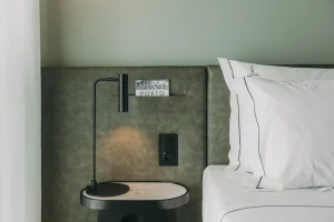 Bedside with a lamp, postcard, and white pillow in a minimalist hotel room.