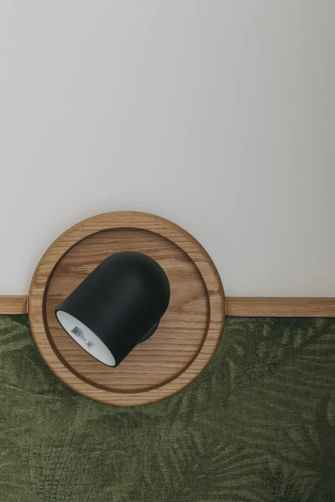 Black spotlight fixture on a round wooden base, mounted on a wall with textured green fabric.