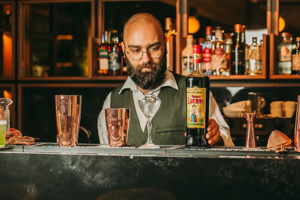 Bartender holding a bottle of Amaro Lucano behind a bar with cocktail tools.